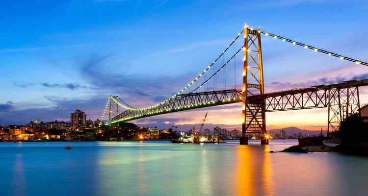 Puente colgante brillando al atardecer sobre aguas tranquilas con cielo pastel y el horizonte de la ciudad de fondo