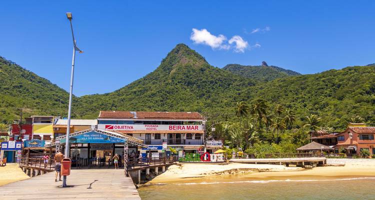 Village côtier coloré avec jetée, commerces et montagnes verdoyantes s'élevant derrière sous un soleil éclatant