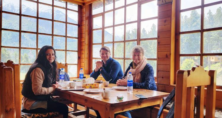 Trois voyageurs savourent le petit-déjeuner dans un restaurant en bois douillet offrant une vue panoramique sur les pics enneigés et la forêt.