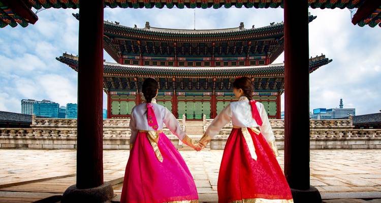 Twee vrouwen in kleurrijke hanbok-jurken houden elkaar vast terwijl ze naar een sierlijke paleisplaats kijken die is ingelijst door een poort.