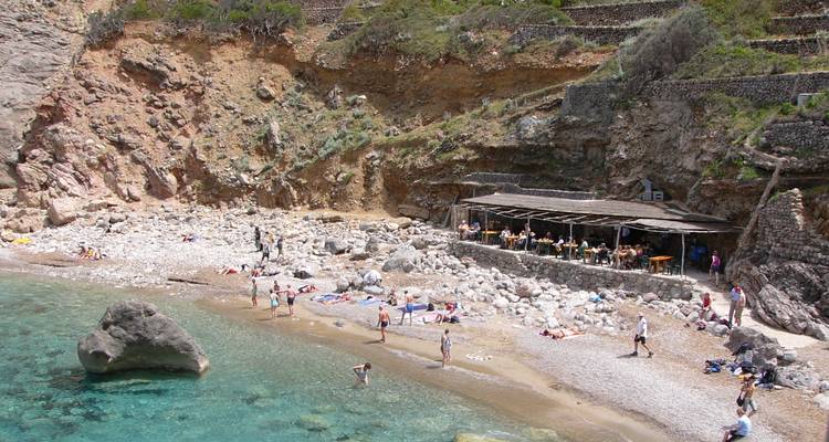 Felsige Bucht mit klarem türkisfarbenem Wasser, wo Badegäste neben einem rustikalen Strandcafé sonnenbaden.