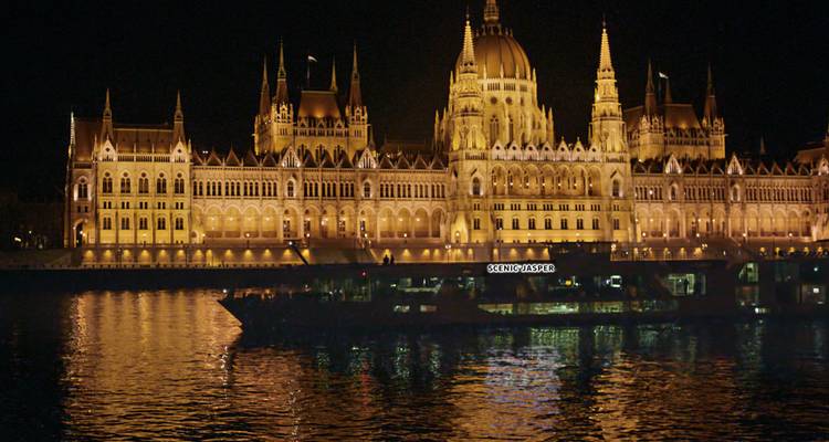 Night view of the illuminated Hungarian Parliament Building with a river cruise boat reflecting on the Danube.