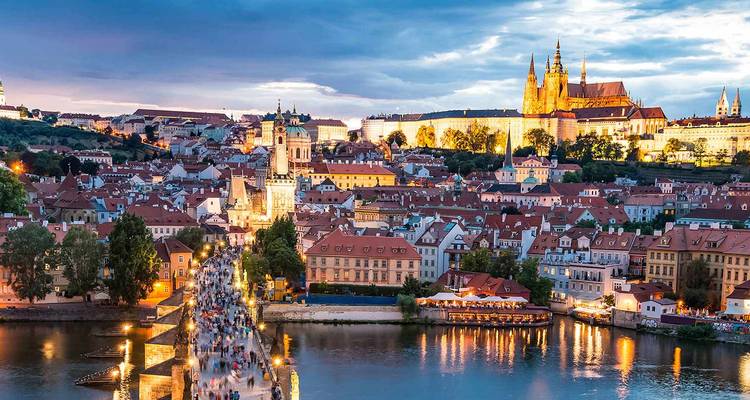 Evening view over Prague with Charles Bridge crowded with people and Prague Castle glowing above the Vltava River.