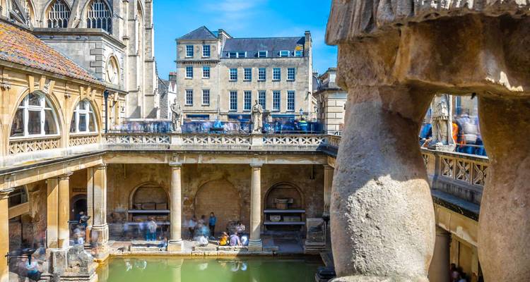 Historic Roman Baths complex in Bath with steaming green pool and stone colonnades under blue sky.