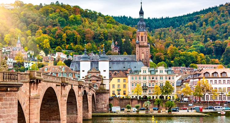 Pont historique en pierre enjambant une rivière menant à la vieille ville de Heidelberg avec clocher d'église et collines boisées aux couleurs automnales