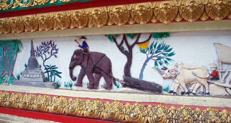 Ein farbenfrohe Flachrelief-Wandmalerei zeigt einen Elefanten und Nutztiere an einer laotischen Tempelwand.