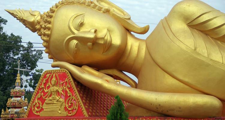 Eine massive liegende goldene Buddha-Statue ruht friedlich im Freien mit dekorativen roten Sockelelementen.