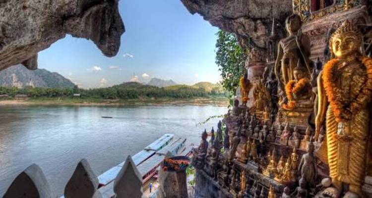 Buddhistische Statuen, die mit Ringelblumengirlanden geschmückt sind, überblicken einen breiten Fluss von innen einer felsigen Höhlenöffnung aus.