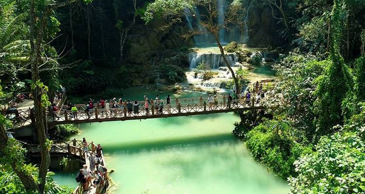 Touristen überqueren eine Holzbrücke über türkisfarbene Pools und Wasserfälle in einem üppigen Tal.