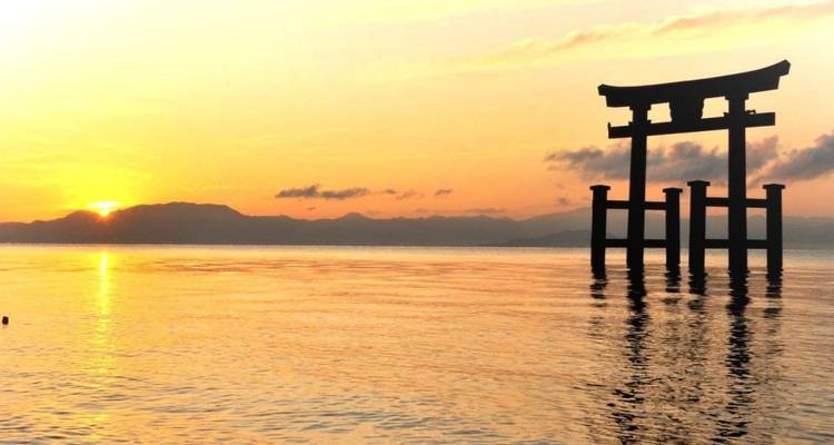 Silhouette of a floating torii gate against an orange sunset over calm water.