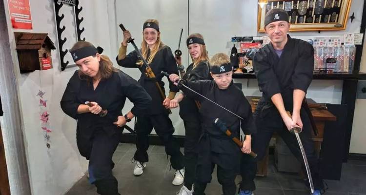 Five visitors dressed in black ninja outfits pose playfully with toy swords inside a studio