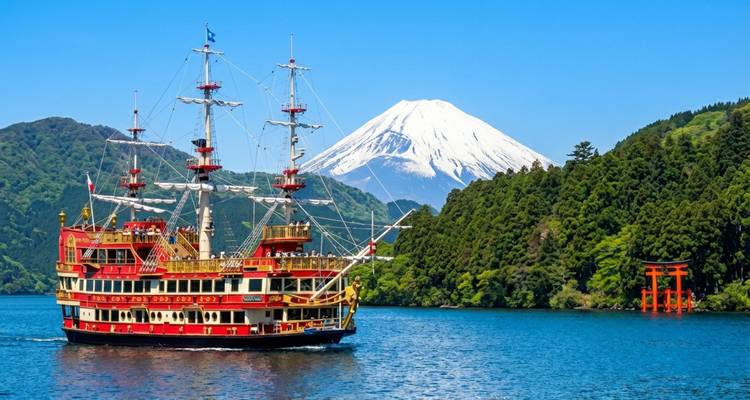 Red-and-gold pirate ship cruises across a blue lake with lush hills, a vermilion torii gate, and snow-capped Mount Fuji in the background