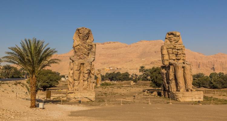 Two massive seated stone statues of the Colossi of Memnon standing before a desert mountain backdrop.