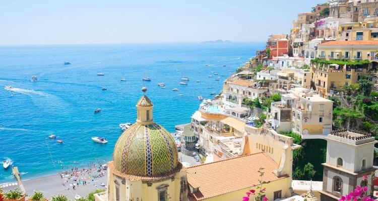 Kleurrijke rij huisjes die langs de rotswand van Positano naar beneden vallen naar een felblauwe zee vol bootjes.