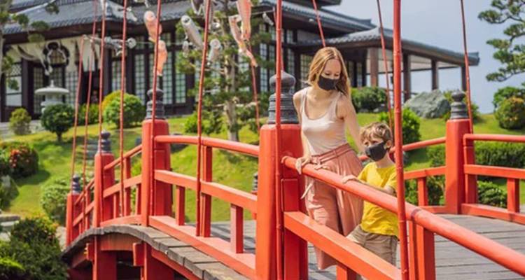 Mother and young son wearing face masks cross a bright red bridge in a manicured Japanese garden.