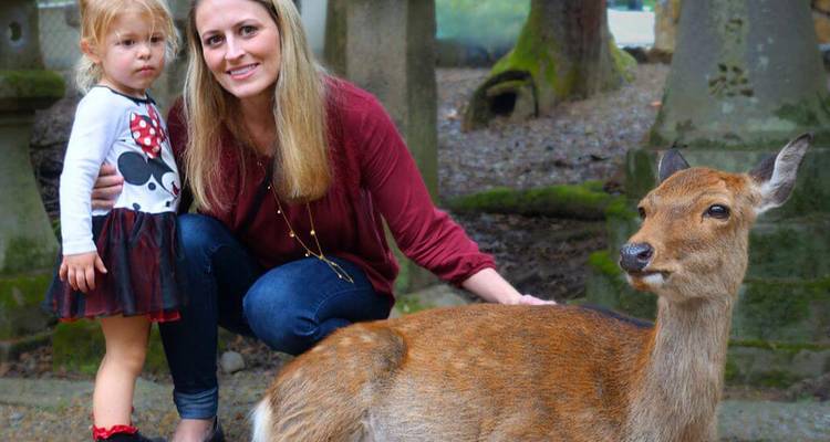 Mother and young daughter interact with a tame deer in a mossy temple setting.