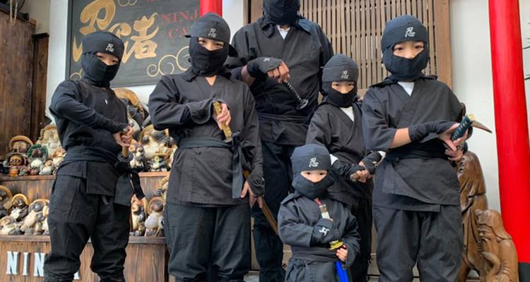 Children and adults dressed in black ninja costumes pose with toy swords outside a themed attraction.