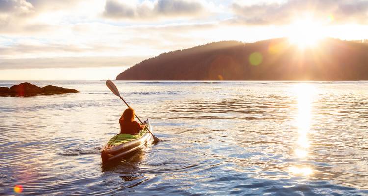 A solo kayaker paddles across calm coastal waters toward the setting sun with a forested headland in the distance.
