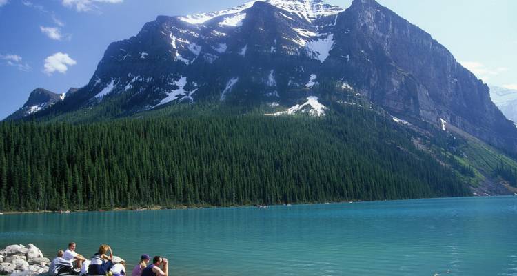 A small group relaxes on the rocky shore of a glacial lake backed by a steep forested mountain face.