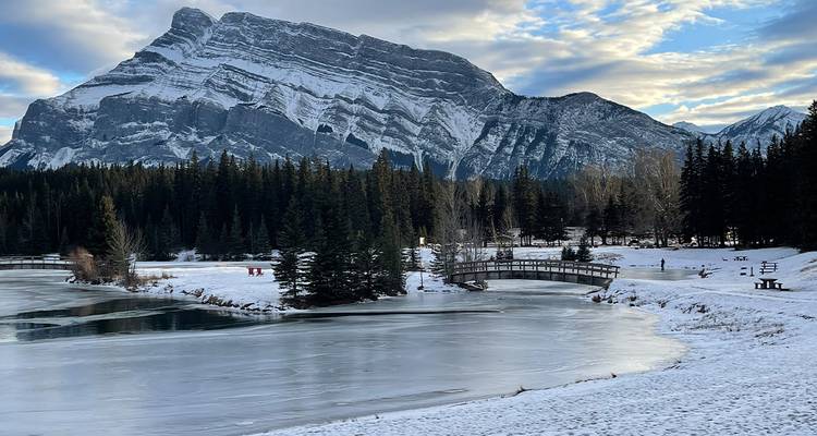 Partially frozen lake reflecting snow-covered Mount Rundle with pine forest and wooden bridge in Banff National Park.