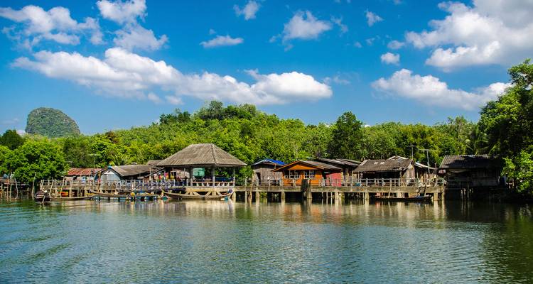 Small riverside village with wooden houses on stilts surrounded by lush forest under blue sky.