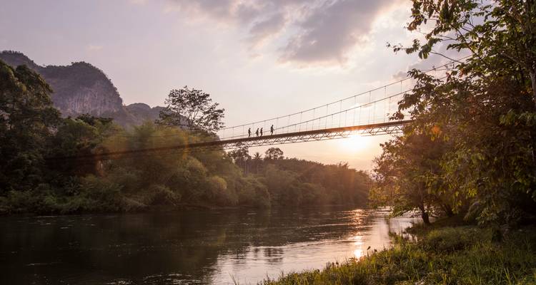 Silhouettes of hikers cross a narrow suspension bridge over a reflective river at sunset.