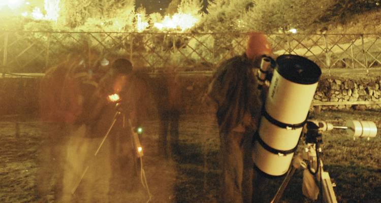 Long-exposure night scene of people using telescopes for stargazing under yellow lights.
