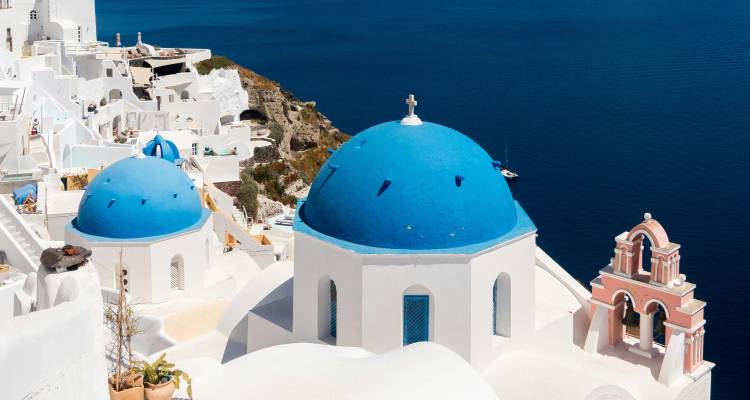 Los icónicos dómulos azules de Oia contrastando fuertemente con el profundo Mar Egeo abajo bajo un cielo brillante.