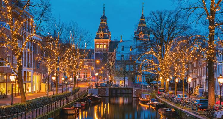 Twinkling holiday lights reflect in a tranquil Amsterdam canal at twilight with historic gabled houses.