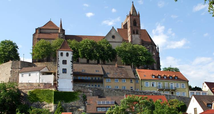 Medieval hilltop church and fortifications rise above a row of traditional houses under blue sky.
