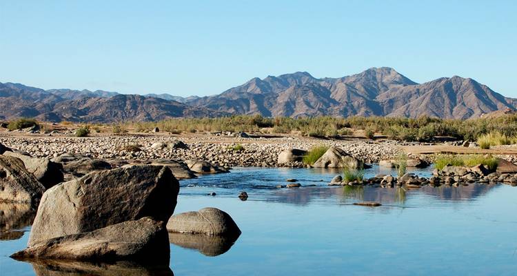 Río tranquilo con rocas dispersas reflejando montañas escarpadas lejanas bajo un cielo azul despejado.