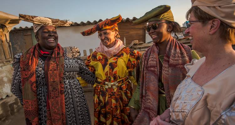 Mujeres sonriendo en coloridos trajes tradicionales namibios compartiendo un momento alegre al atardecer.