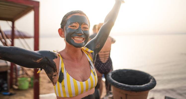 Smiling woman covered in Dead Sea mud raises her arms by the water's edge.