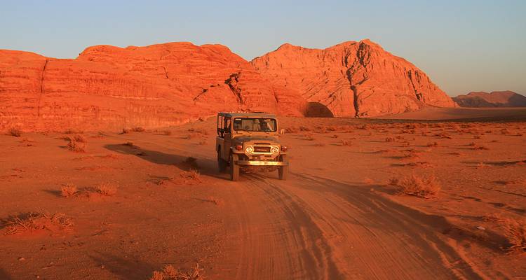 4x4 jeep drives across red sand between towering sandstone cliffs at sunset in Wadi Rum.