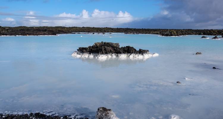 Lagon géothermique bleu laiteux avec des roches de lave noire et des nuages lointains.