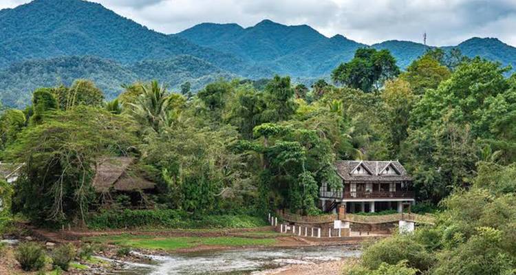 Houten huis aan de rivier verscholen tussen dichte tropische begroeiing met bergen op de achtergrond