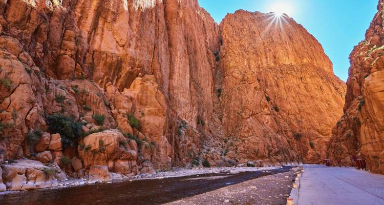 Les imposantes parois de calcaire orange des Gorges du Todra avec un ruisseau qui coule en bas et la lumière du soleil qui apparaît au-dessus de la crête.