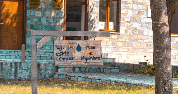 Cartel de madera en albanés instando a la conservación del agua fuera de un edificio de piedra.