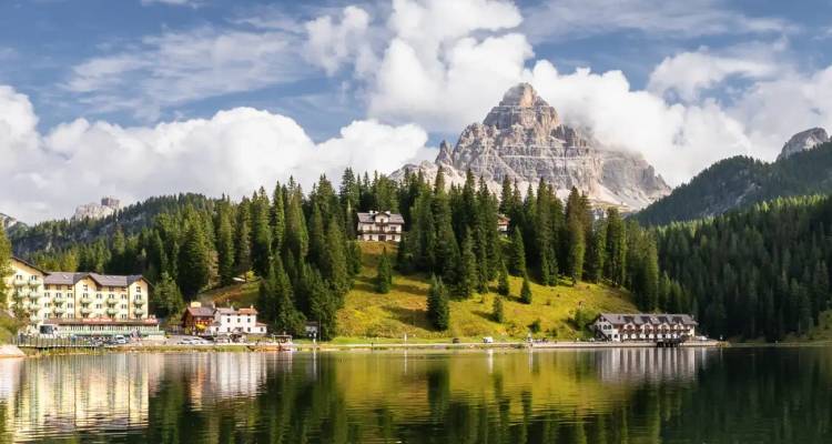 Alpine lake reflecting forested hills, alpine hotels and dramatic Dolomite peak under blue sky.