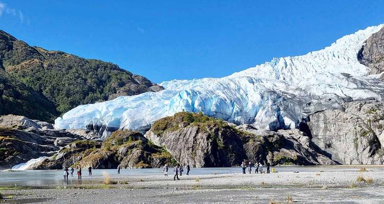 Kleine Figuren erkunden den Fuß eines leuchtend blauen Gletschers, der von zerklüfteten Bergen unter einem klaren Himmel herabfließt.