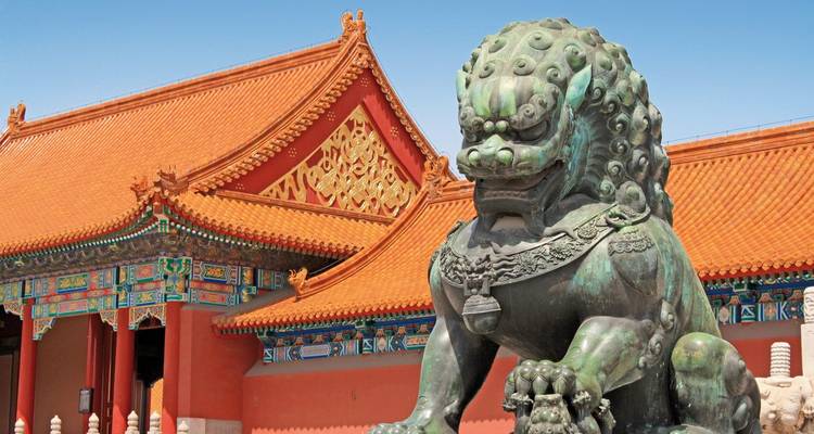 Bronzene Wächterlöwenstatue vor kunstvoll gestalteten roten und goldenen Palastdächern in Beijings Verbotener Stadt unter einem klaren blauen Himmel.