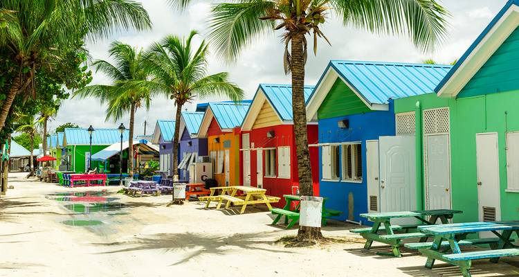 Row of brightly painted beach shacks with palm trees and picnic tables along a sandy Caribbean lane.