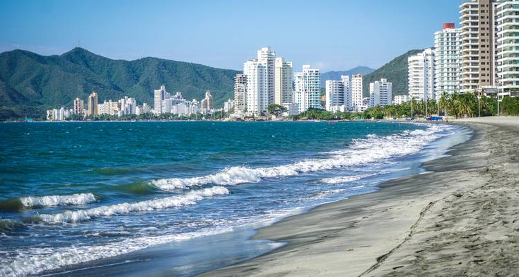Waves crash onto a sandy Caribbean beach with a backdrop of Santa Marta’s modern high-rise skyline.