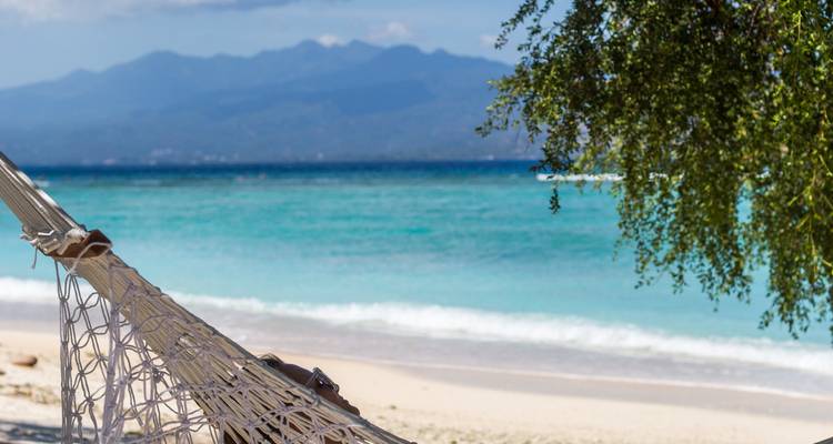 Une personne se détend dans un hamac en filet sur une plage de sable blanc avec vue sur une eau turquoise éclatante et des montagnes lointaines.