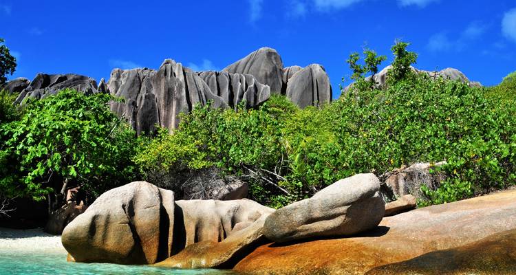 Dramatic granite rock formations and dense greenery rising behind shallow turquoise water.