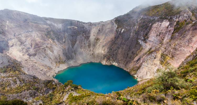 Cratère volcanique aux hauts murs contenant un lac turquoise éclatant partiellement voilé par la brume.