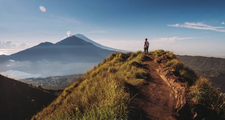 Hiker standing on a grassy ridge overlooking Mount Batur and a volcano-dotted horizon.