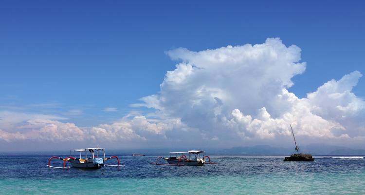 Traditional outrigger boats floating on calm, crystal-clear sea beneath a vast cloudscape.