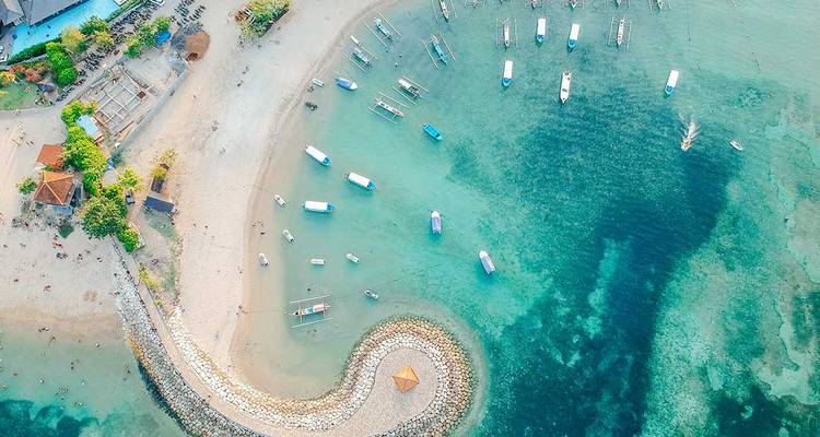 Vista aérea de una playa de arena curva con agua turquesa y pequeñas barcas ancladas en alta mar, bordeada por un rompeolas y vegetación tropical.