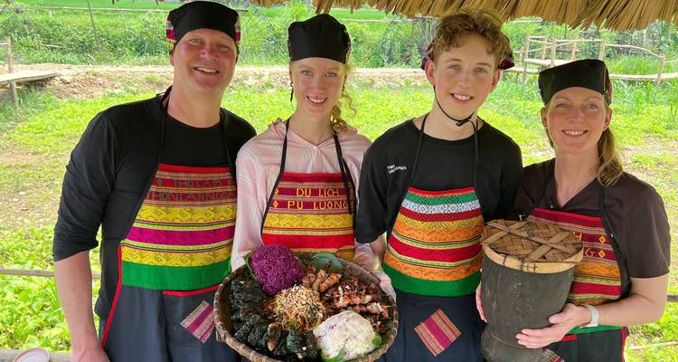 Smiling family wearing colorful aprons presents a platter of local dishes in an open-air rural kitchen.